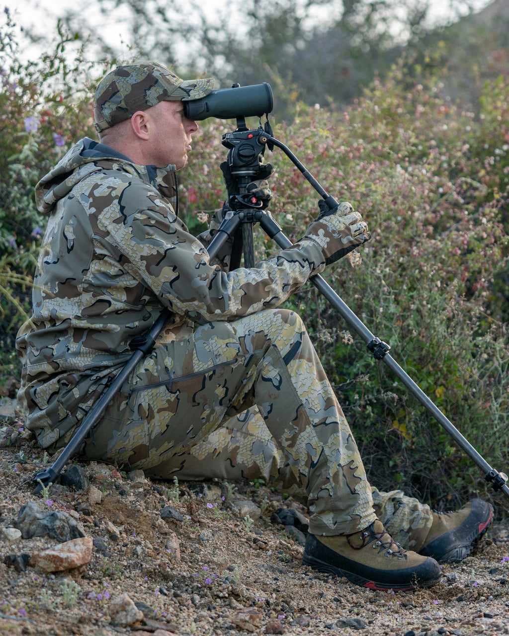 Hunter wearing Kutana Stretch Woven Pant, sitting on sloped, rocky ground, looking through his spotting scope mounted on a tripod