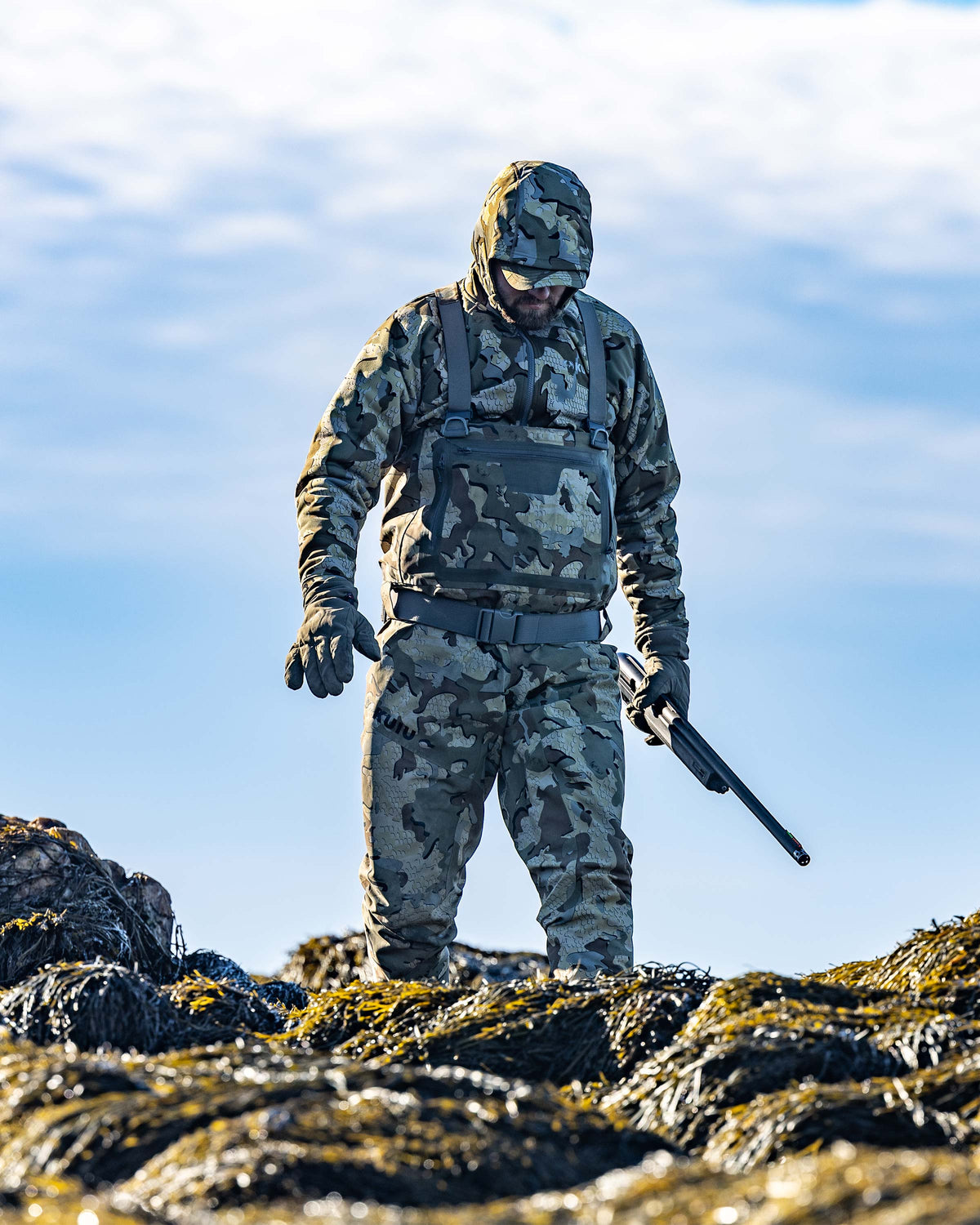 Hunter Wearing HD Flex Waders in Valo Camouflage Holding a Shotgun on Rocky Beach with seaweed on the rocks