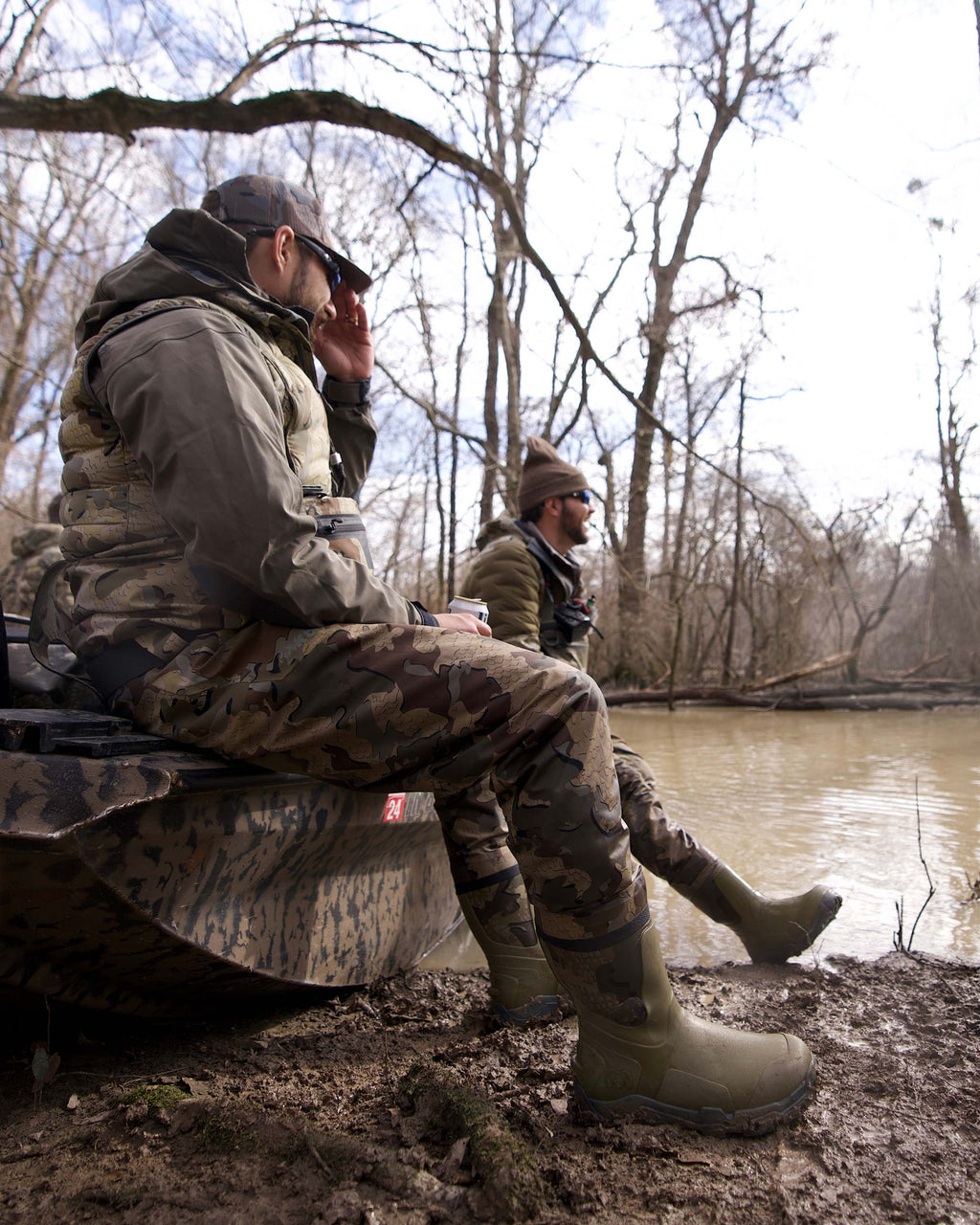 Two Hunters wearing HD Flex Waders in Valo Camouflage sitting on a Duck Boat on the Bank Highlighting the Boot and Self-cleaning Soles