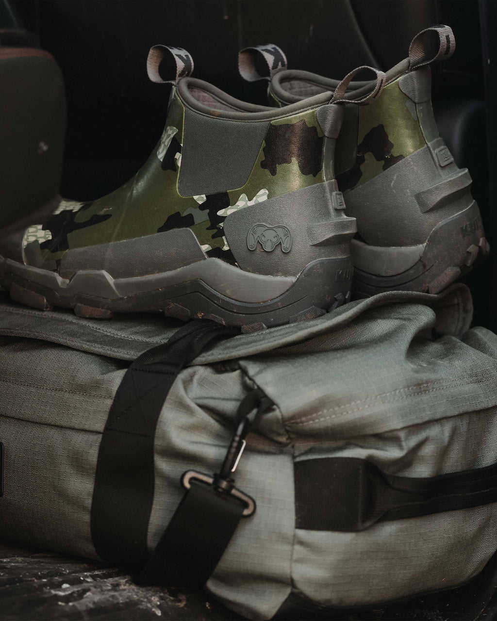 A pair of KUIU Ankle Mud Boot in Verde Camouflage placed on top of a duffel bag, Displaying the side tread boot design and kick-off heel tab 