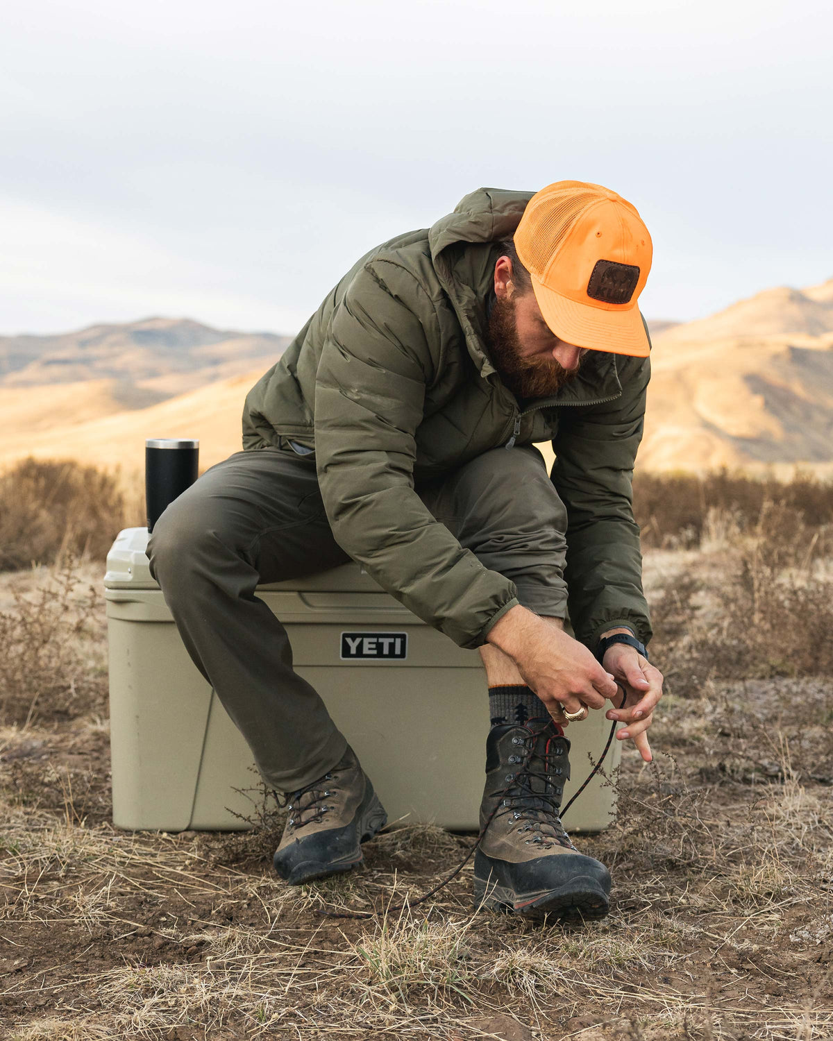Hunter wearing Elements Jacket in Ash Brown tying his boot lace while sitting on a YETI cooler