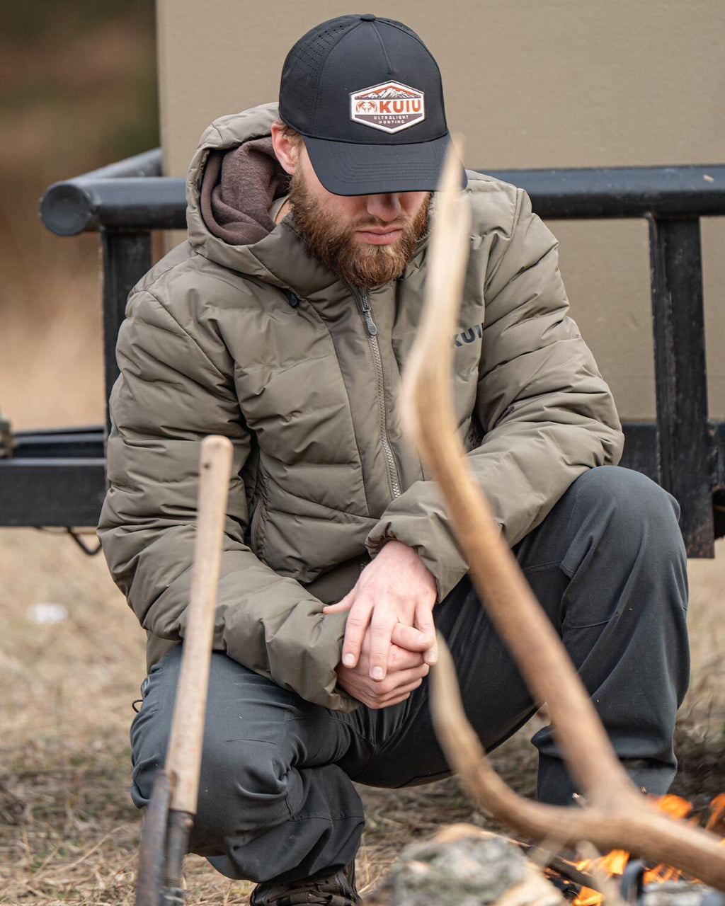 Hunter squatting next to a fire pit, wearing Elements Jacket in Ash Brown