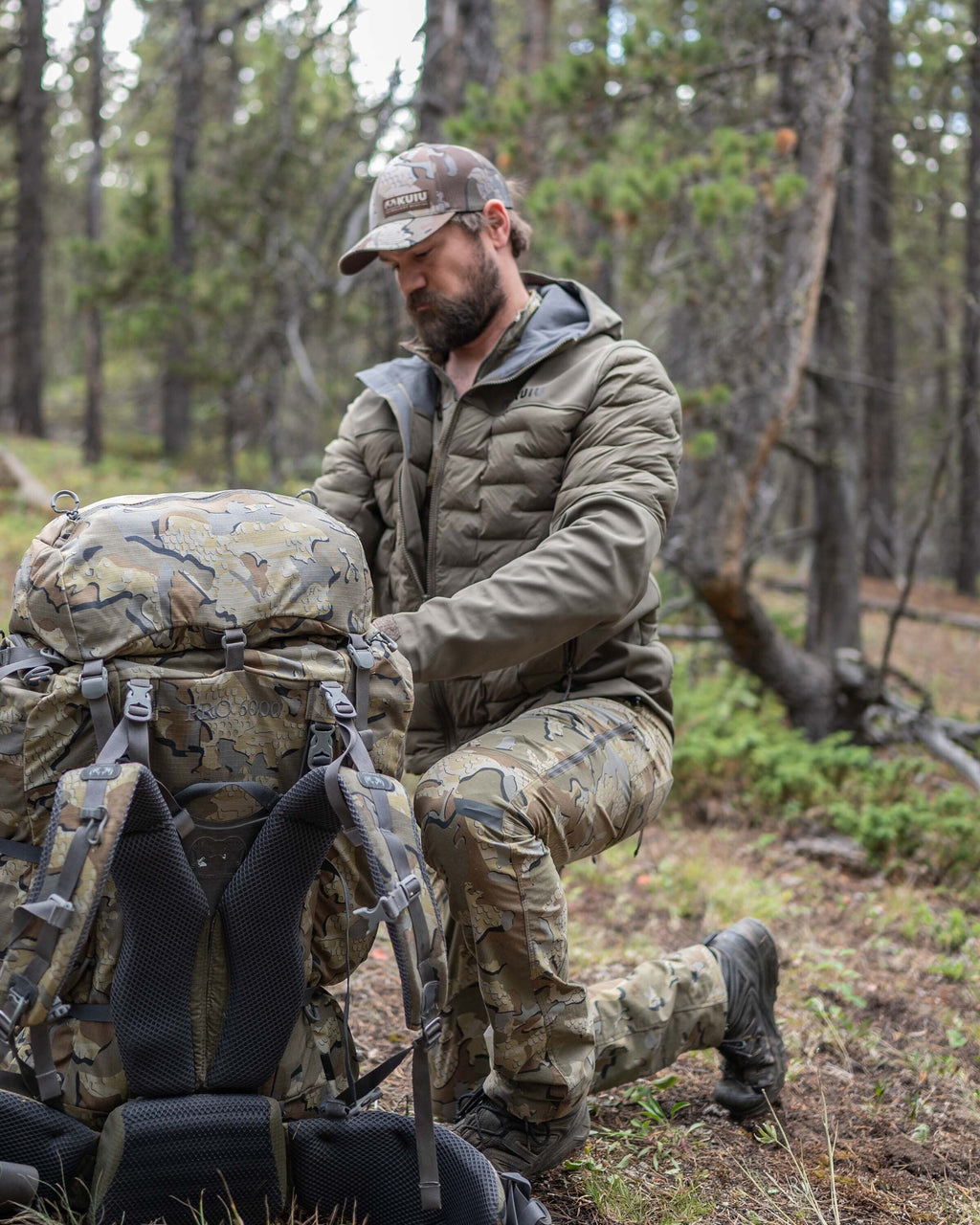 Hunter in wooded area kneeling with pack wearing Axis Thermal Hooded Jacket partially unzipped in Ash Brown