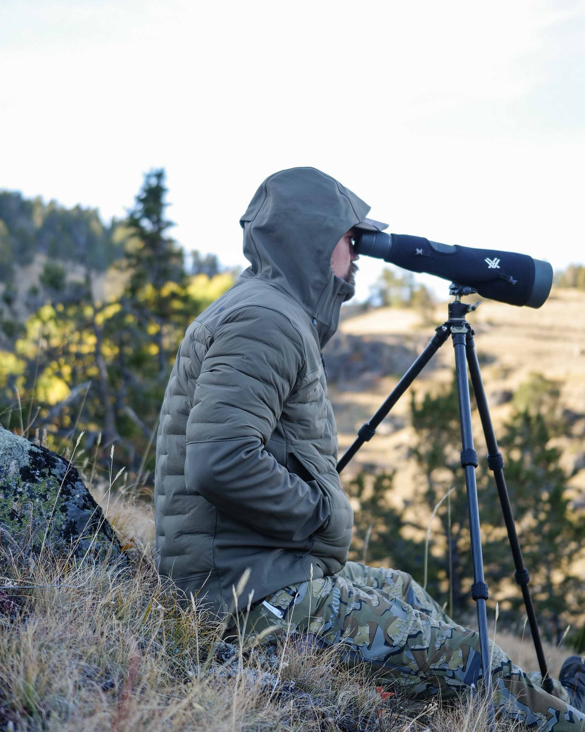 Hunter sitting on hill glassing wearing Axis Thermal Hooded Jacket in Ash Brown