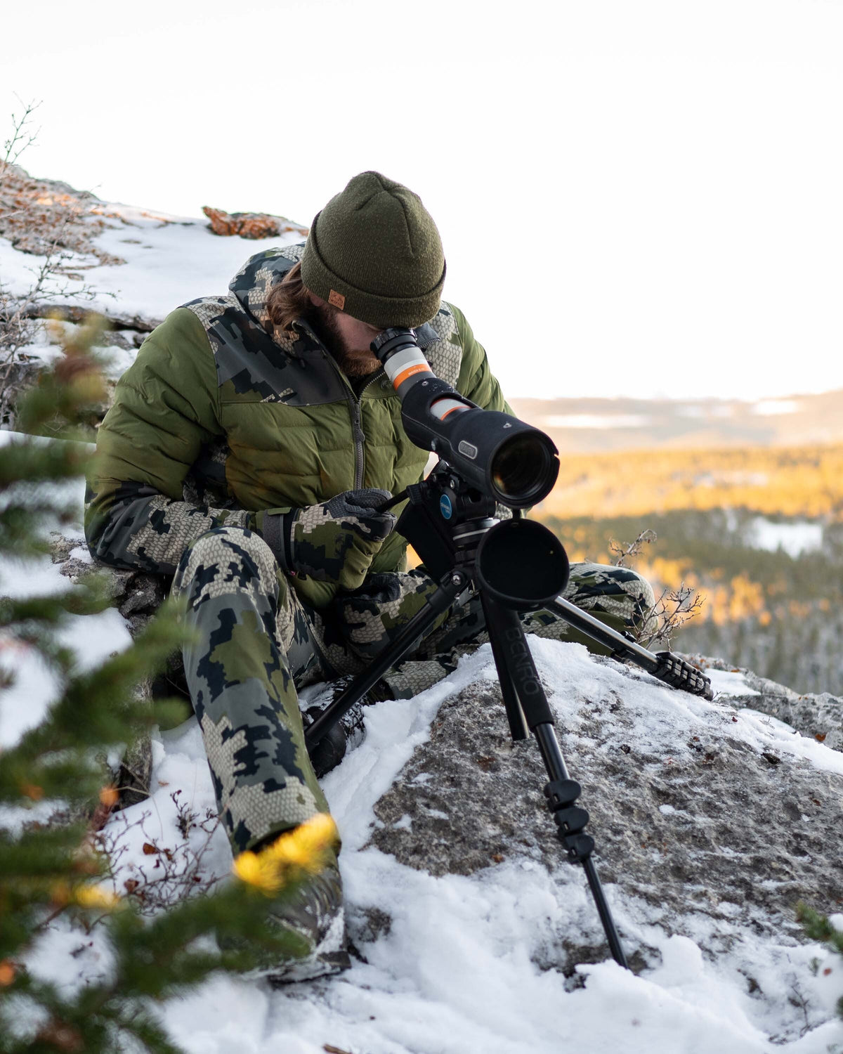 Front, Angled View of Hunter Glassing Game Through Spotting Scope Wearing Axis Thermal Hybrid Hooded Jacket in Verde Camouflage