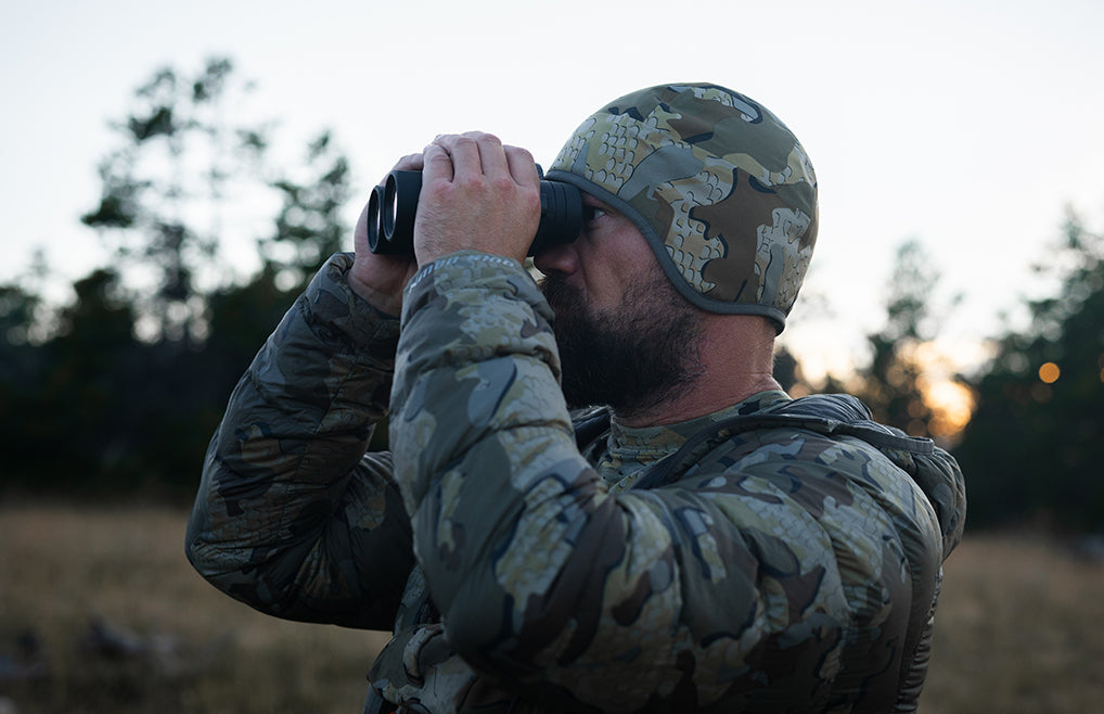 A man in a camouflage hunting jacket and matching beanie looks through binoculars outdoors in a field at sunset.