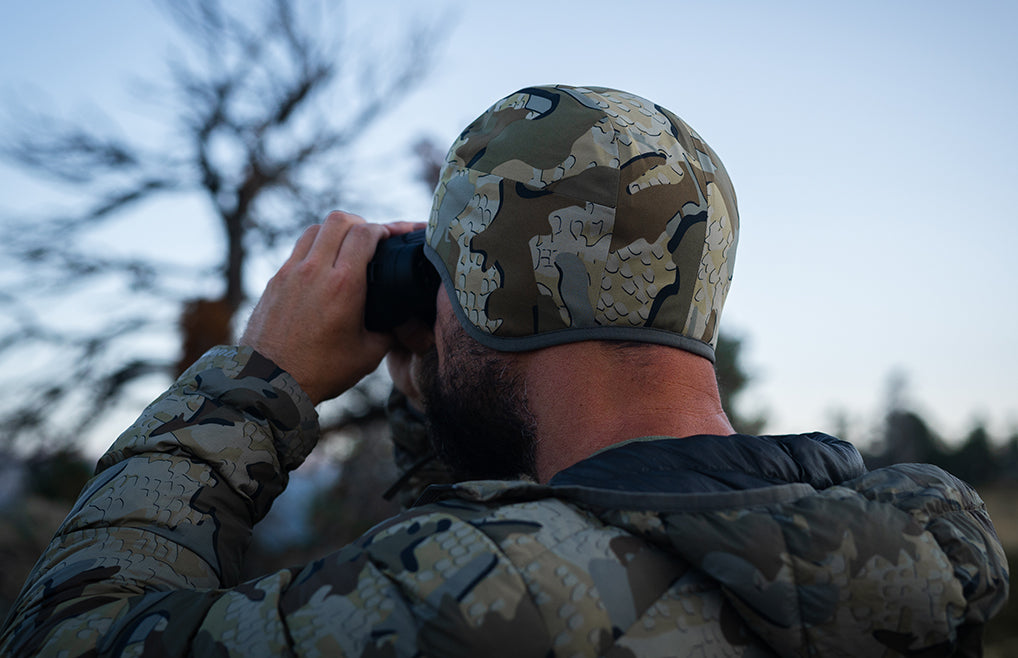 Hunter wearing a Mountain PRO Beanie in Valo Camouflage and matching camouflage jacket, seen from behind as he looks through binoculars outdoors.
