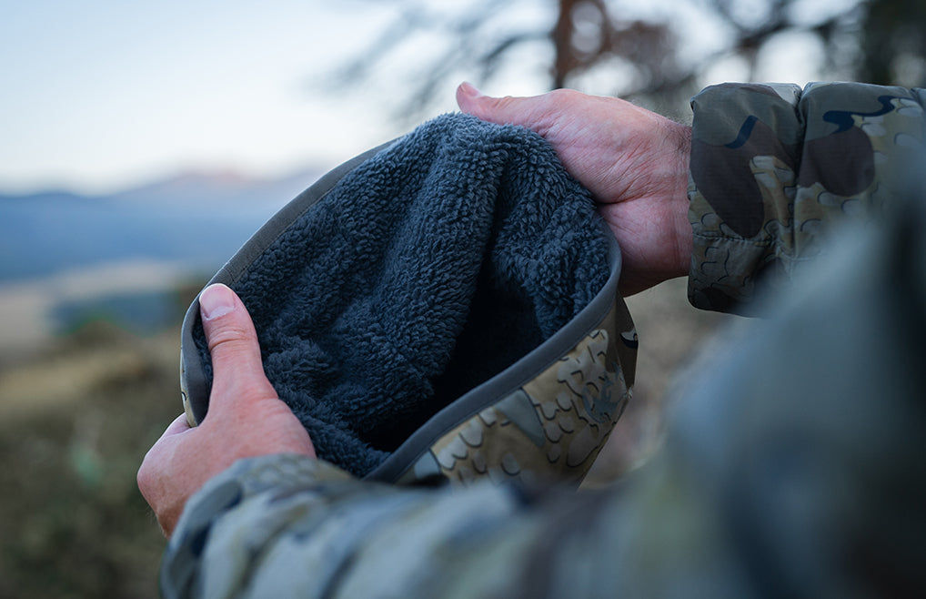 A close-up of a person holding Mountain PRO Beanie in Valo Camouflage, Highlighting fleece-lined beanie with Valo camouflage-patterned exterior, set against a blurred outdoor mountain background.