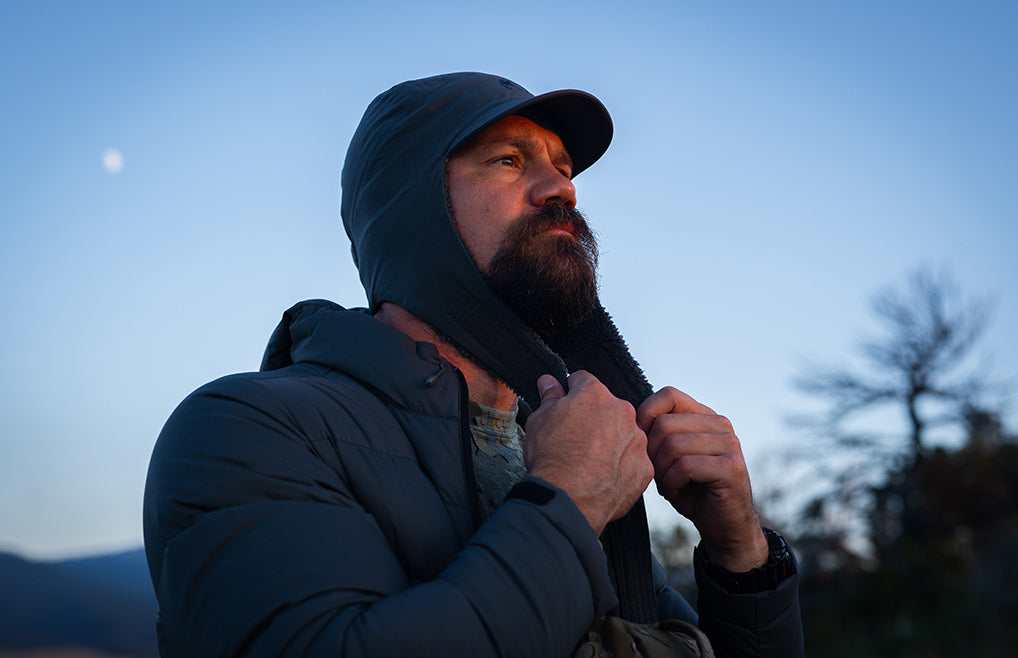 A man wearing a Mountain PRO Hat in Gunmetal Grey pulling down on the hook-an-loop connection strap, looking upward outdoors against a blue twilight sky.