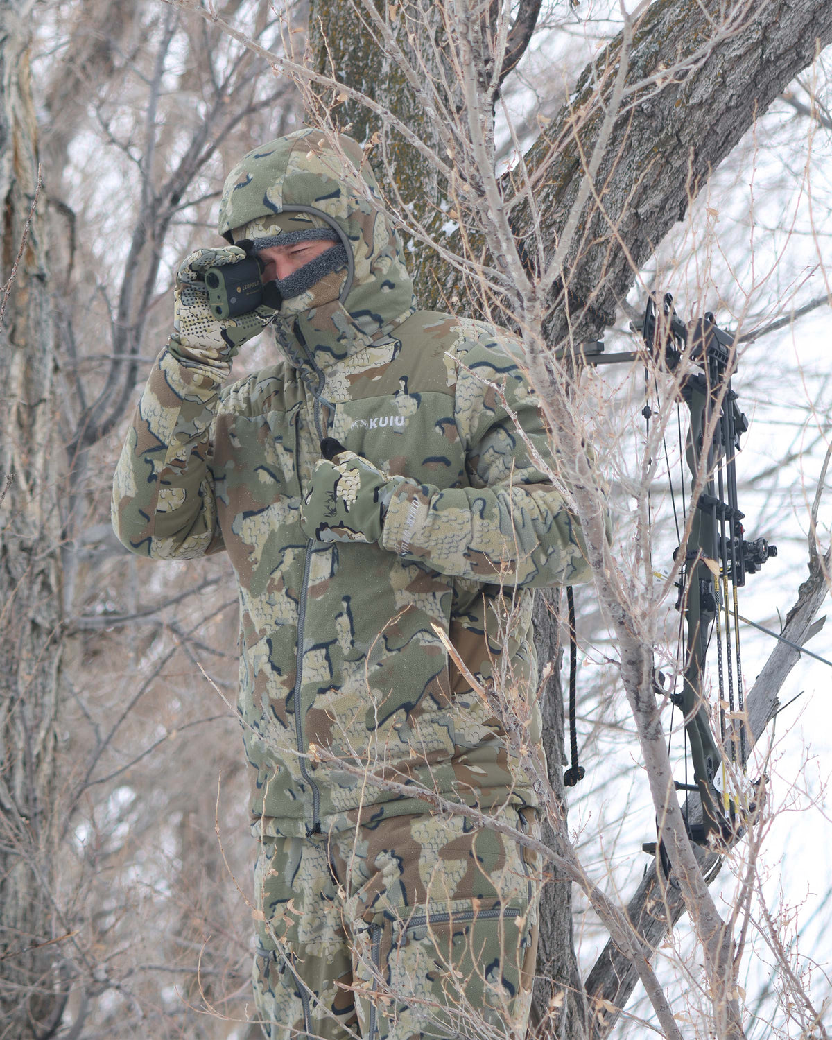 Hunter standing camouflaged in a tree stand, glassing, wearing Super Down Haven Hooded Jacket in Valo Camouflage