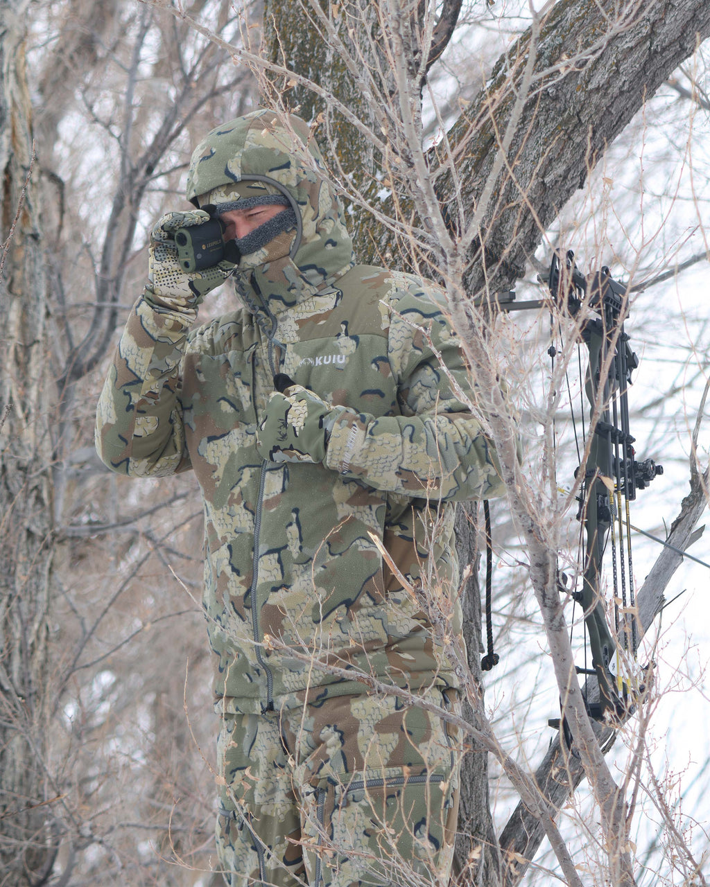 Hunter standing camouflaged in a tree stand, glassing, wearing Super Down Haven Hooded Jacket in Valo Camouflage