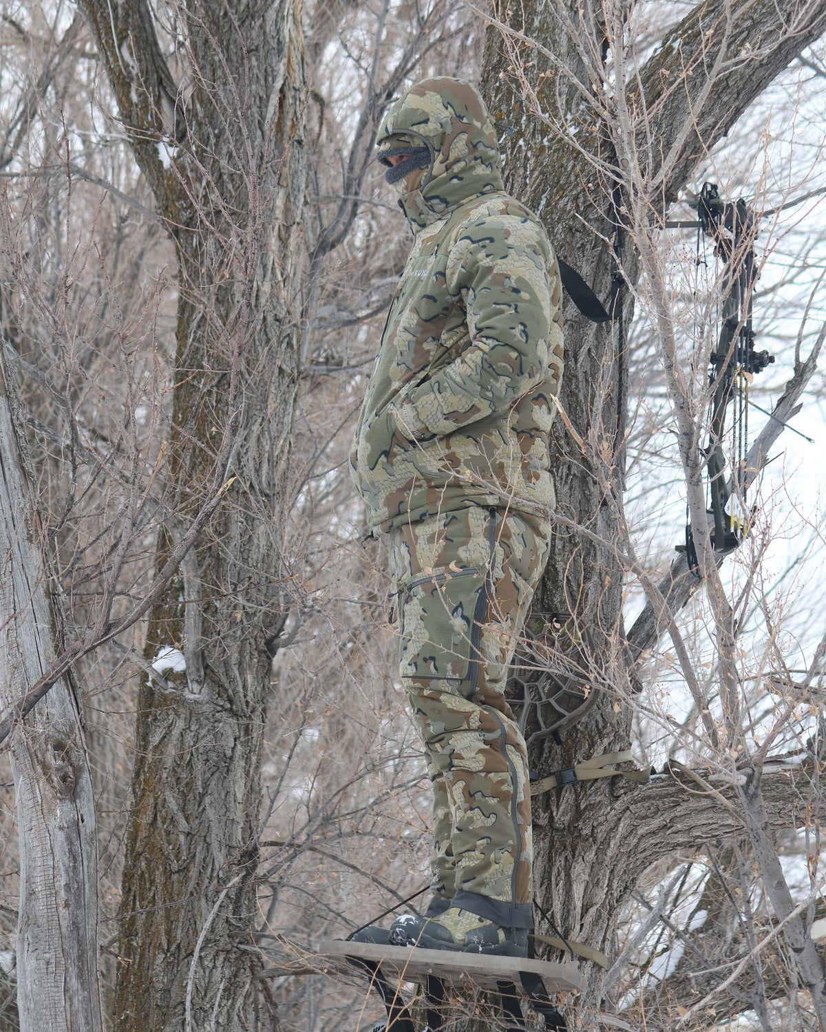 Hunter standing camouflaged in a tree stand with hands in his pocket, wearing Super Down Haven Hooded Jacket in Valo Camouflage