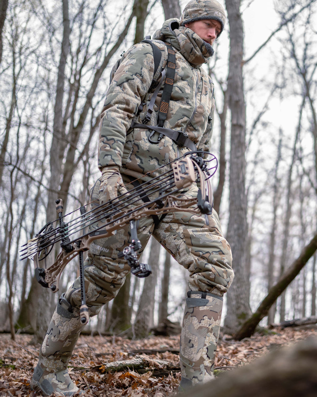 Hunter hiking through the woods, carrying a bow, wearing Super Down Haven Hooded Jacket in Valo Camouflage