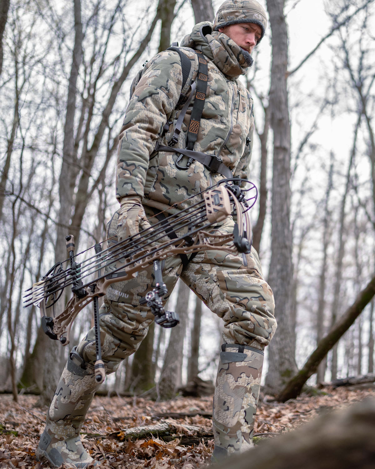 Hunter hiking through the woods, carrying a bow, wearing Super Down Haven Hooded Jacket in Valo Camouflage