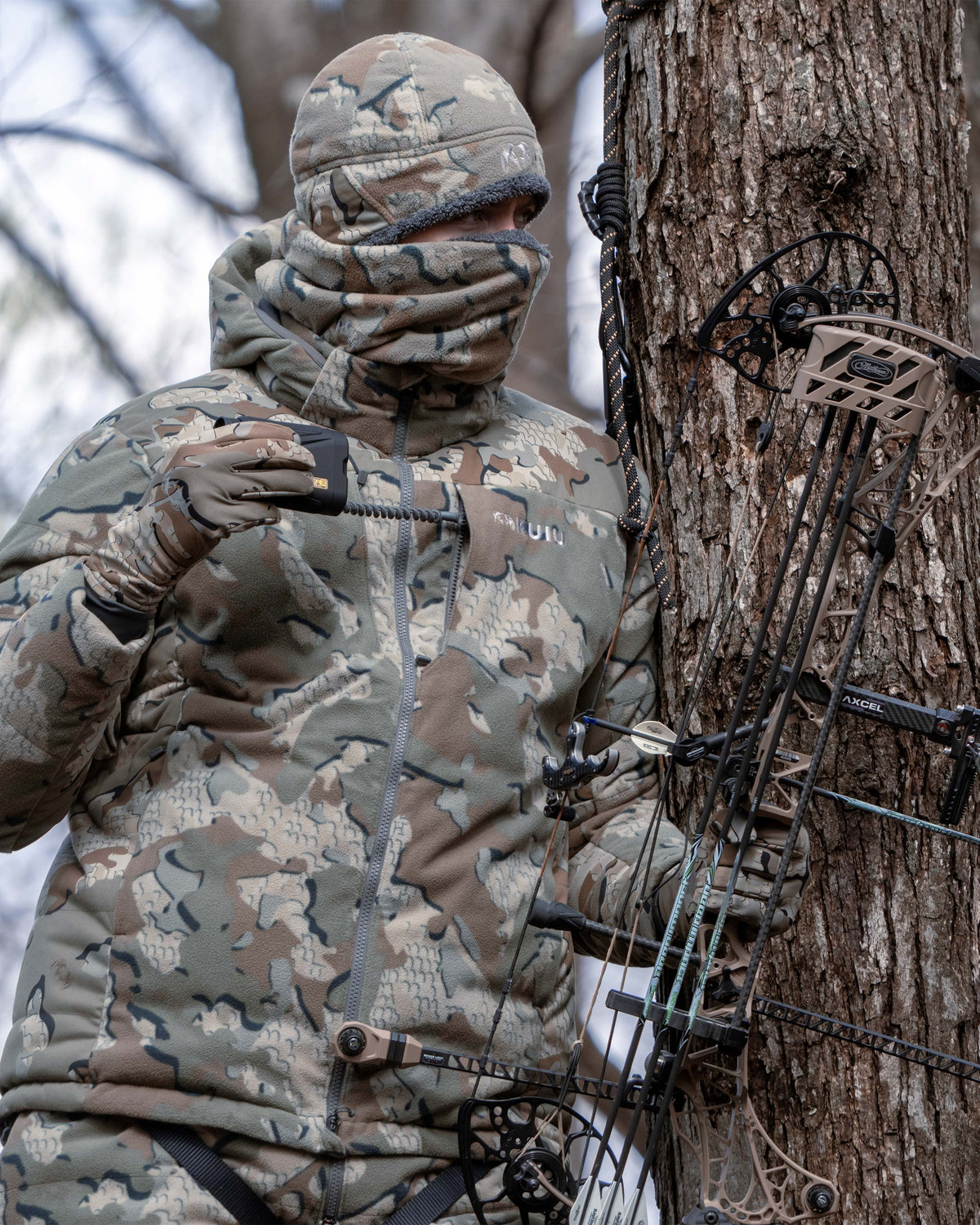 Hunter in a tree stand holding a bow and arrow, wearing Super Down Haven Hooded Jacket in Valo Camouflage