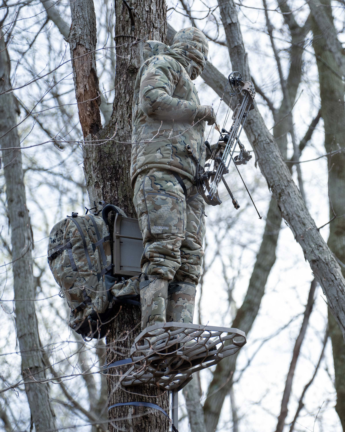 Hunter camouflaged in a tree stand, holding a bow and arrow, wearing Super Down Haven Hooded Jacket in Valo Camouflage