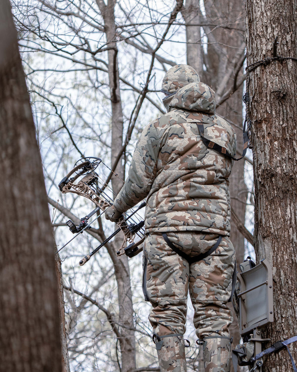 Back view of Hunter in a tree stand, holding a bow and arrow, blending into the forest surroundings, wearing Super Down Haven Hooded Jacket in Valo Camouflage