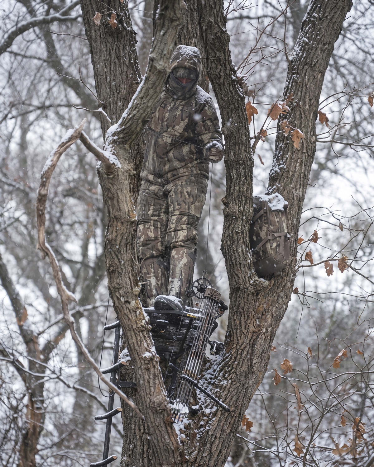 Whitefowl hunter a Super Down Haven Bib  in Veros camouflage stands in a tree stand attached to a tree trunk in a snowy forest.