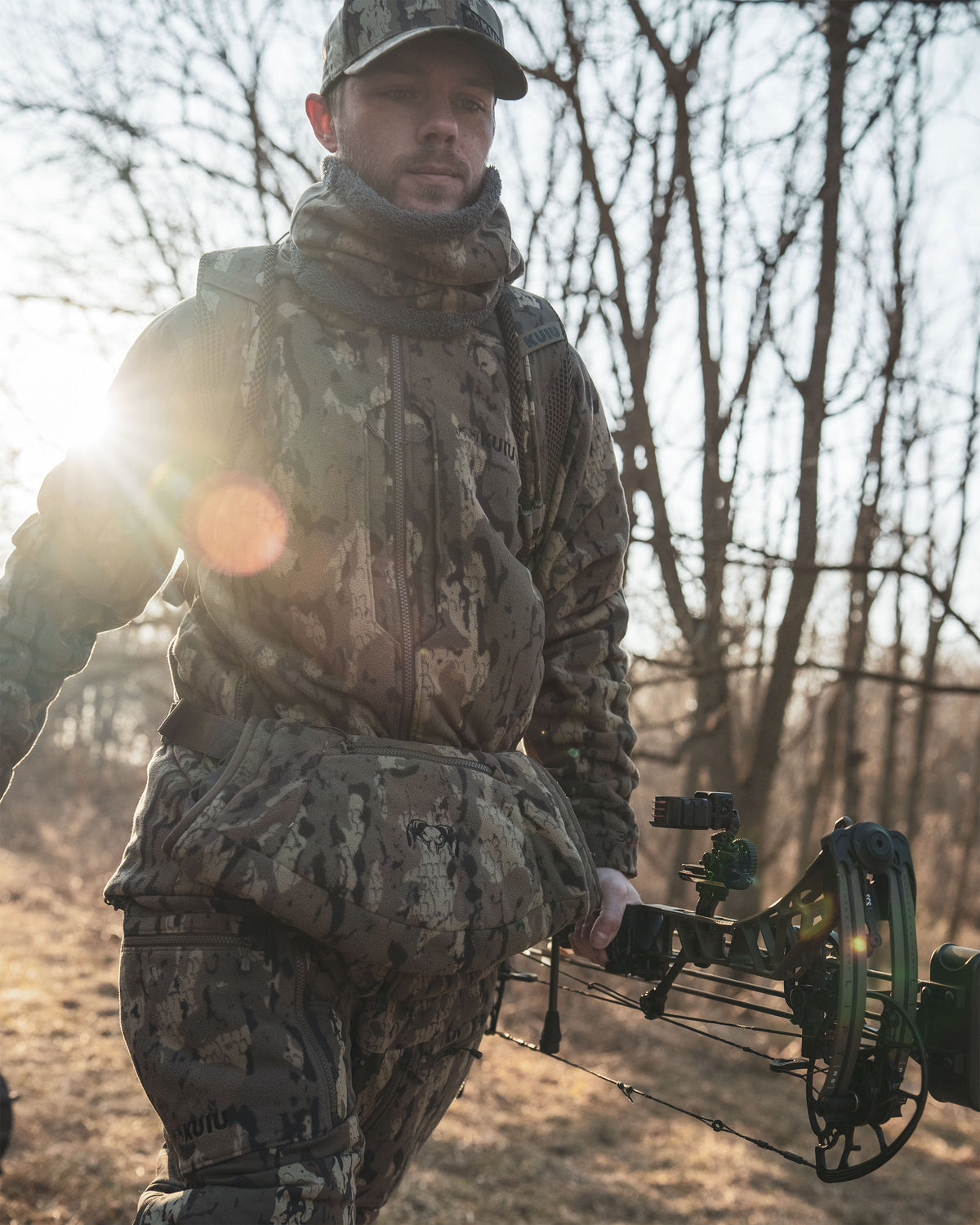 Hunter in full Veros camouflage hunting apparel and Proximity Hand Muff stands outdoors in a wooded area at sunrise, holding a compound bow. The image features strong backlighting and lens flare.