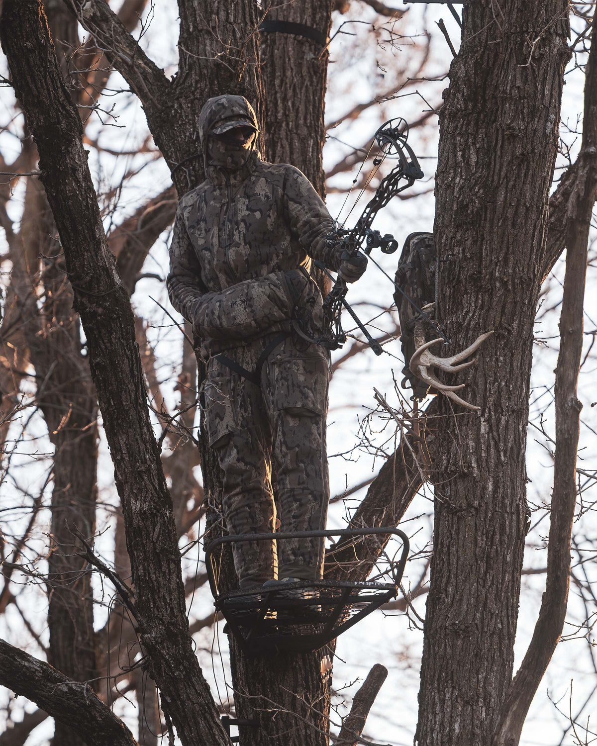 Hunter in full KUIU Veros Camouflage clothing and Proximity Hand Muff in Veros Camouflage stands in a tree stand holding a compound bow, surrounded by bare trees in a forest setting.