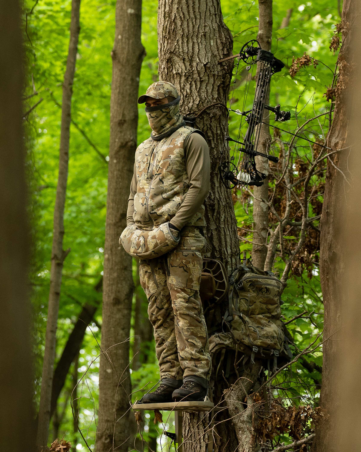 Hunter Using Proximity Hand Muff in Valo Camouflage Standing in a Treestand in Wooded Area with Pack and Bow