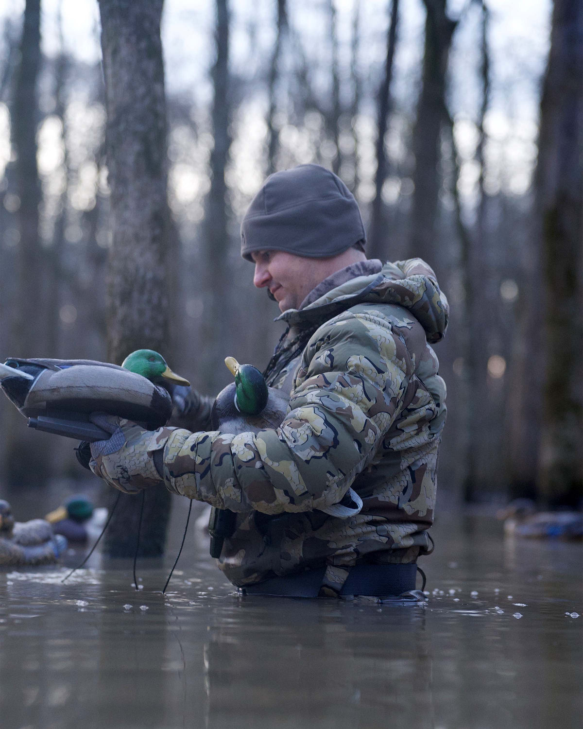 Side of Hunter Wearing Flyway Insulated Hooded Jacket in Valo Camouflage Holding Drake Mallard Decoys in Flooded Timber