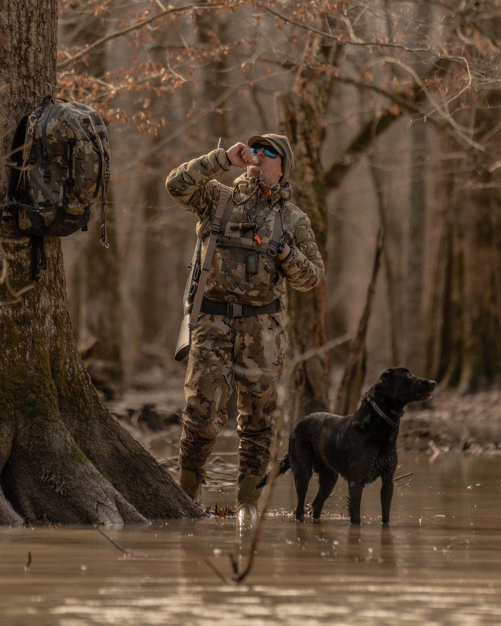 Hunter Wearing Flyway Insulated Hooded Jacket in Valo Camouflage Calling Ducks in Flooded Timber with Black Labrador
