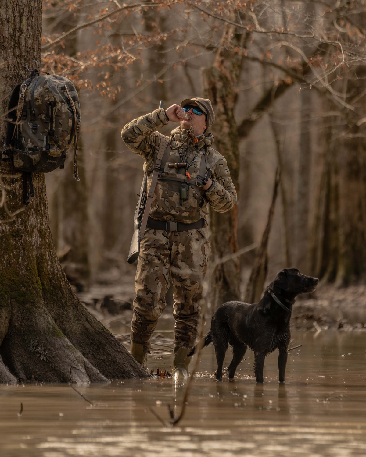 Hunter Wearing Flyway Insulated Hooded Jacket in Valo Camouflage Calling Ducks in Flooded Timber with Black Labrador