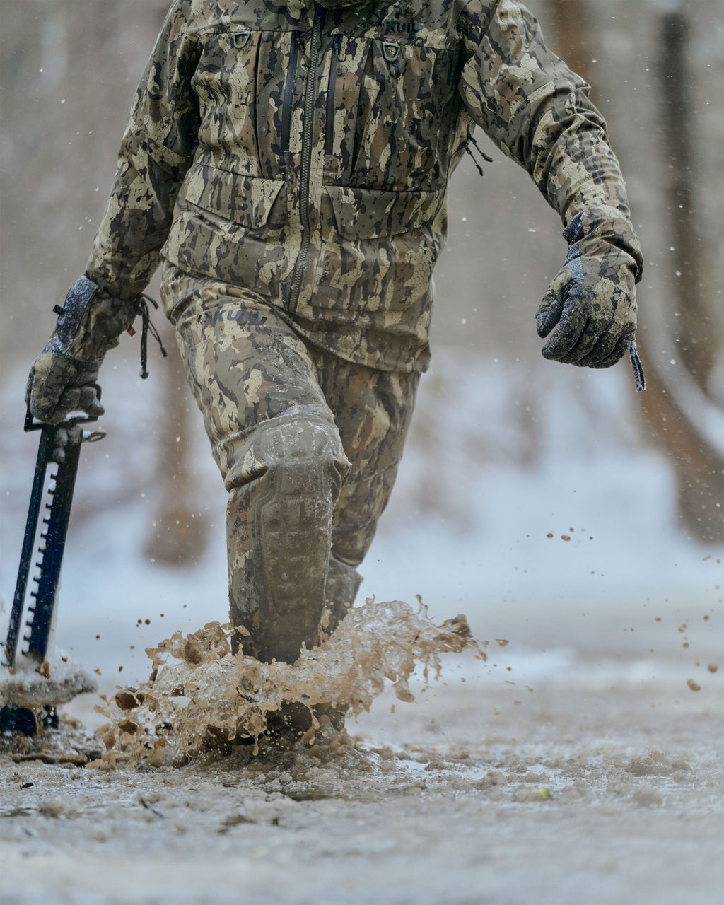 Hunter walks through deep mud and splashing water outdoors in a snowy environment wearing HD Flex Storm Shell Jacket and PRO Hd Flex Zip Wader in Veros Camouflage.