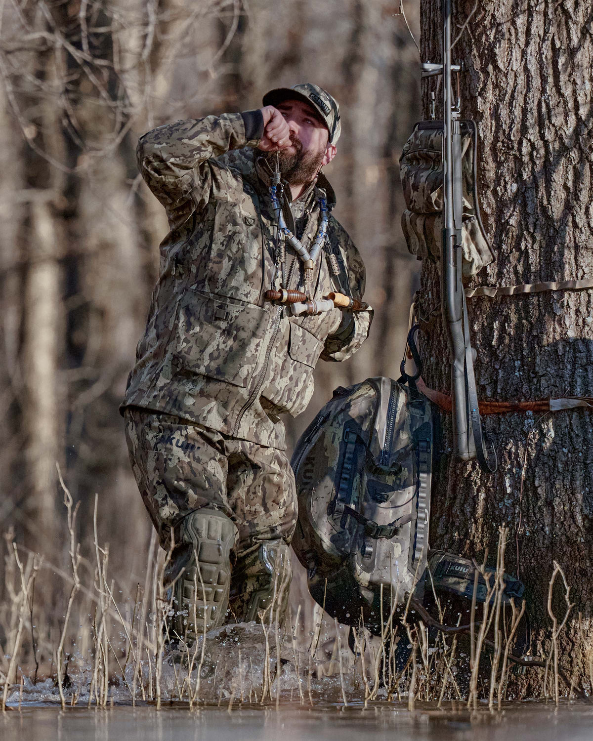 Waterfowl hunter in a KUIU PRO HD Flex Zip Wader and HD Flex Storm Shell Jacket in Veros camouflage stands in shallow water near a large tree, blowing a duck call in a wooded wetland.