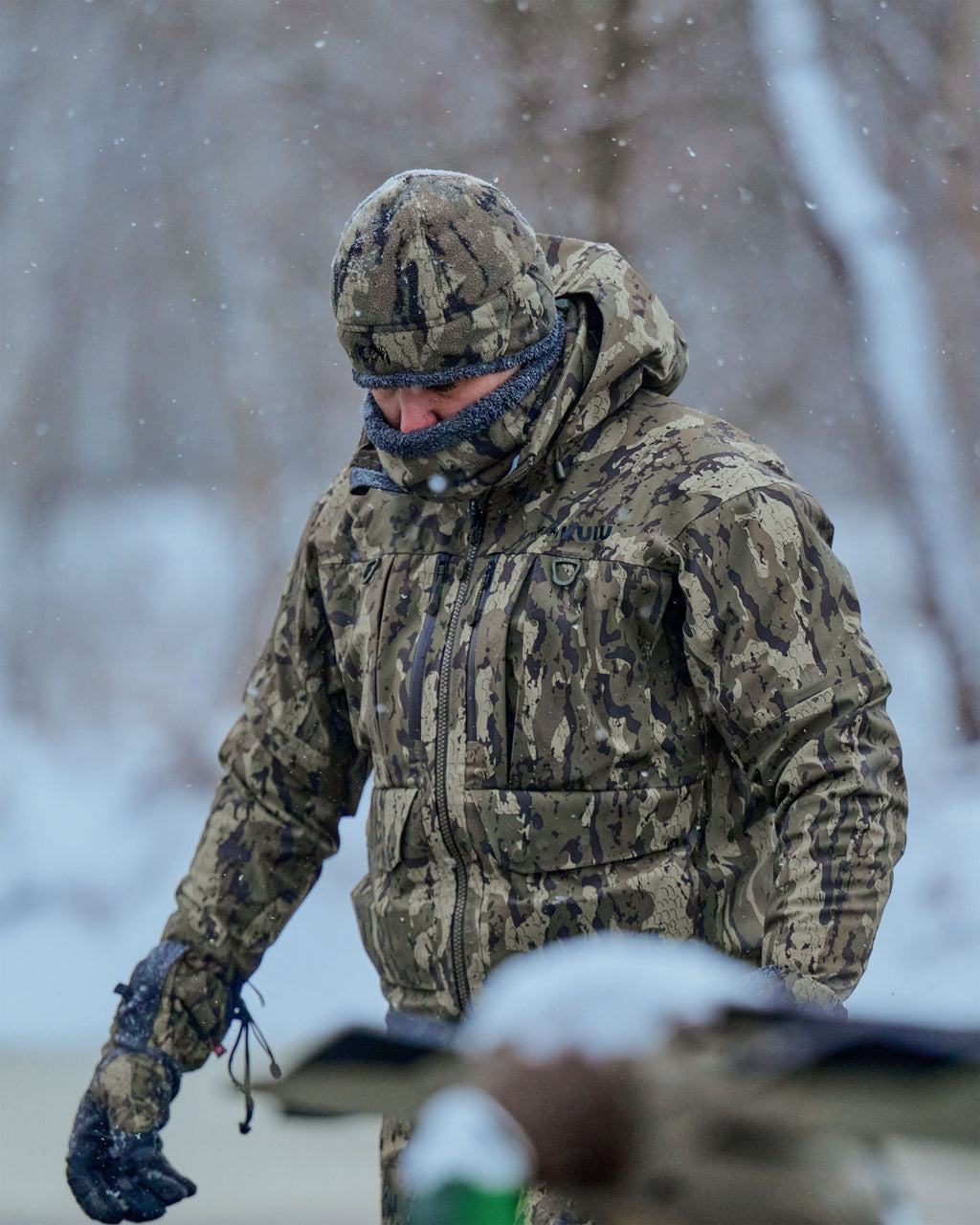 Hunter wearing a HD Flex Storm Shell Jacket, Neck Gaiter and Beanie in Veros Camouflage walks through falling snow outdoors.