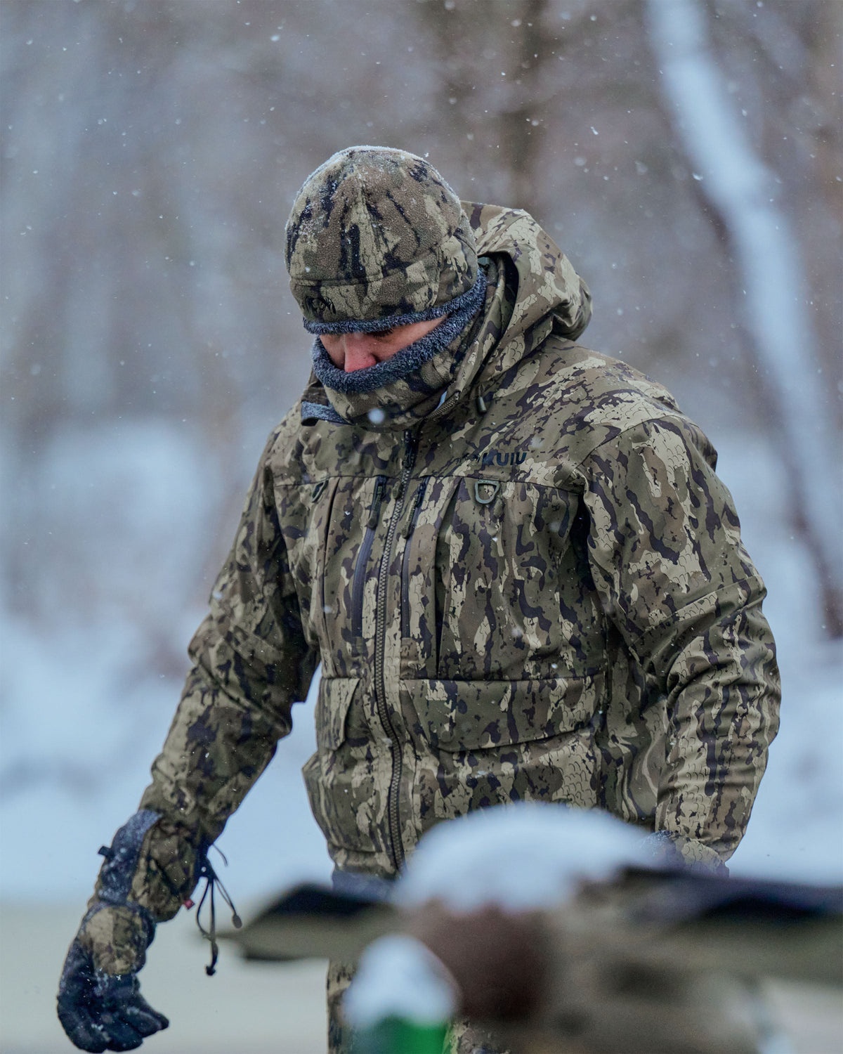 Hunter wearing a HD Flex Storm Shell Jacket, Neck Gaiter and Beanie in Veros Camouflage walks through falling snow outdoors.