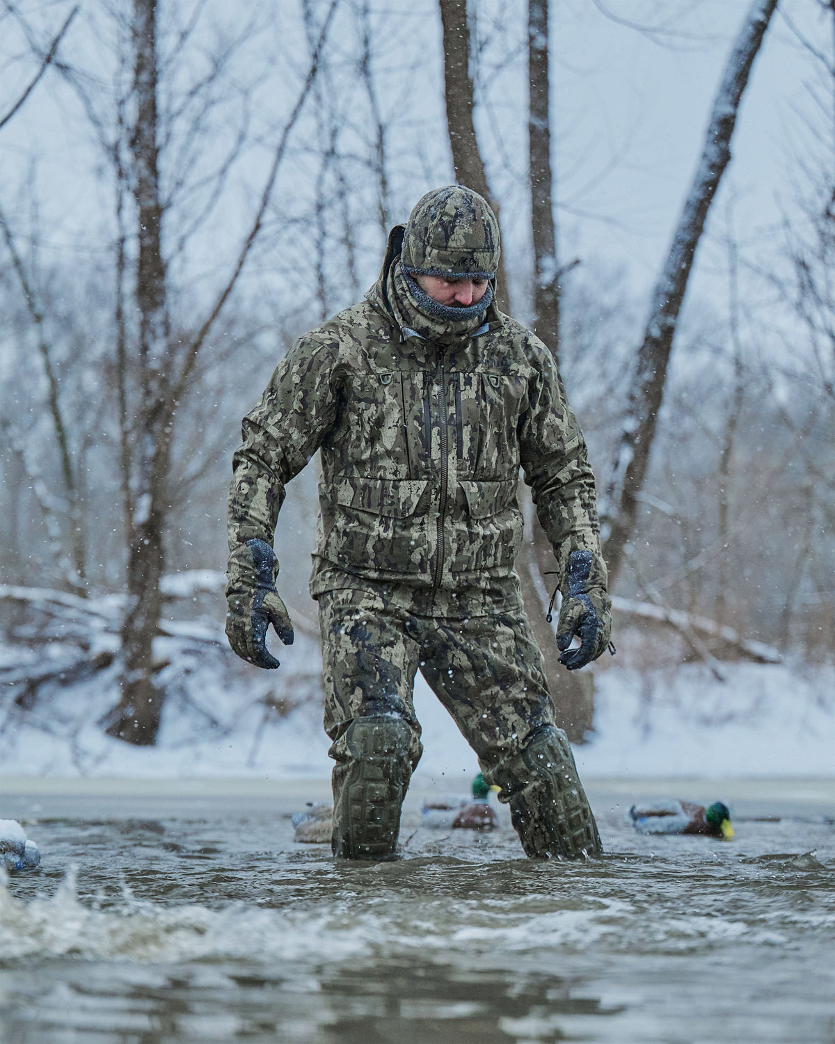 Hunter wearing HD Flex Storm Shell Jacket in Veros Camouflage including beanie and gloves, wading through shallow water in a snowy outdoor setting with bare trees in the background.