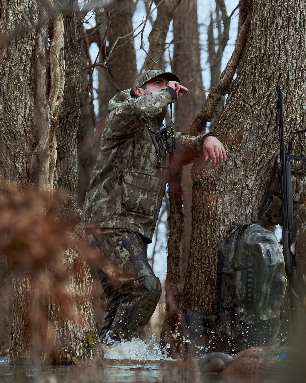 Waterfowl hunter wearing HD Flex Storm Shell Jacket in Veros Camouflage stands in shallow water between large trees, blowing a duck call. A DeltaStorm 2200 Submersible Backpack in Veros Camouflage rests against a tree on the right, with bare winter trees and branches in the background.