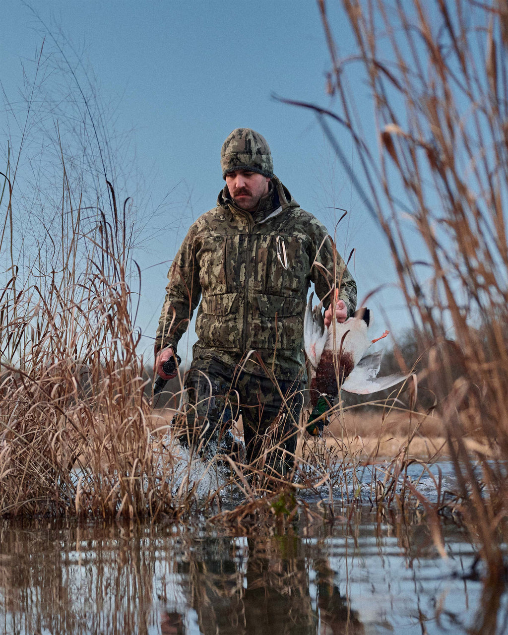 Waterfowl hunter wearing HD Flex Storm Shell Jacket in Veros Camouflage stands in shallow water among reeds, holding a harvested duck. The scene is outdoors in a marsh under a clear blue sky.