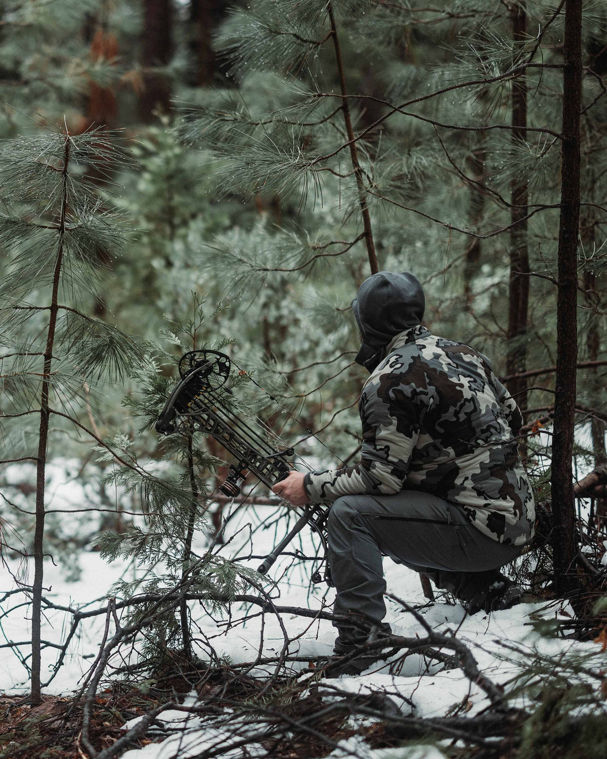 Angled, Rear View of Hunter Sitting in Pine Forest Wearing PRO Brush Pant in Gunmetal Grey Color