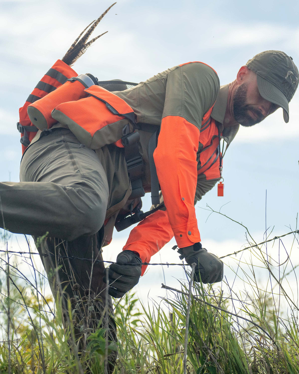 Hunter stepping over a single barb wired fence, wearing Upland Sporter Shirt in Khaki/Blaze Orange