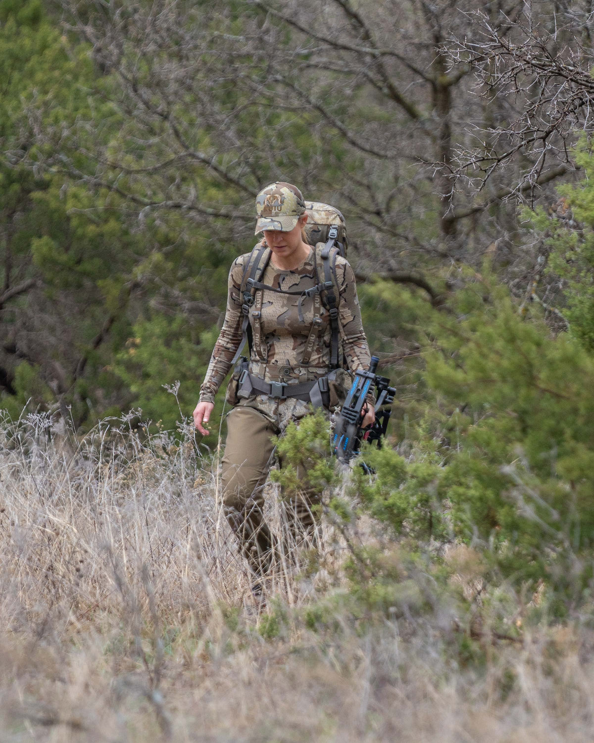 Woman hunter walking through brush terrain carrying bow in one hand wearing Women's PRO Brush Pant in Valo Camouflage