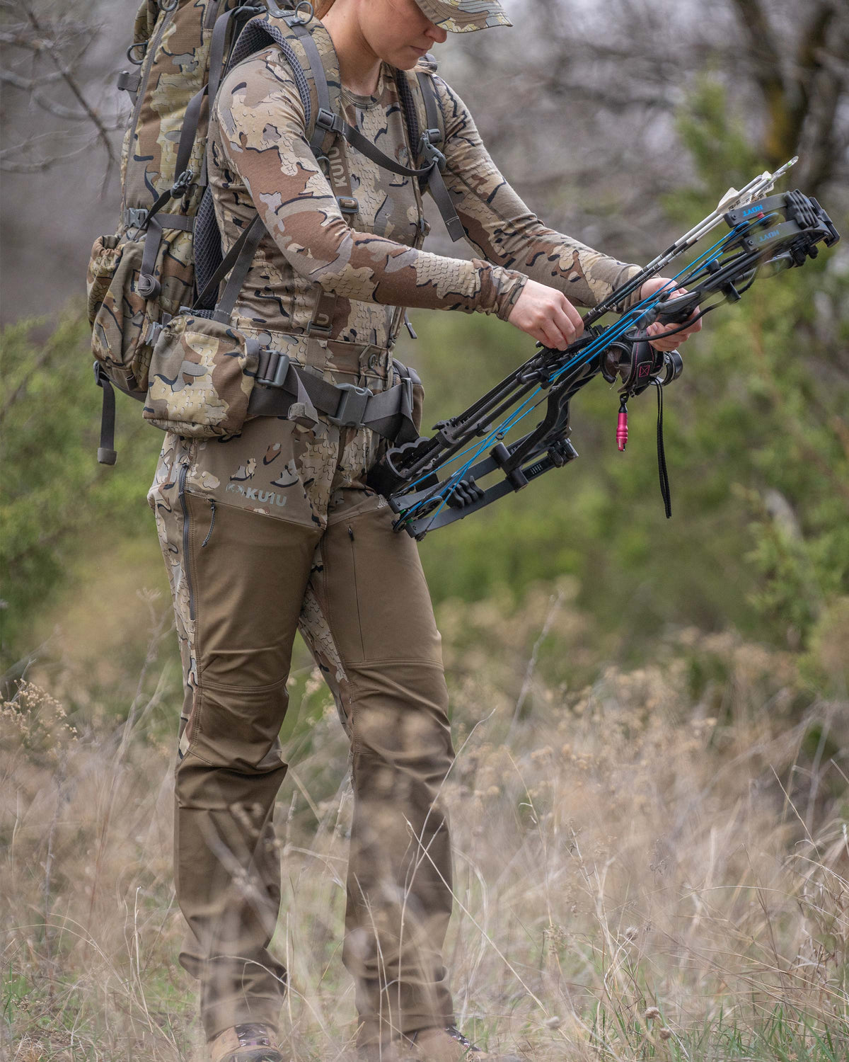 Woman hunter standing in brush terrain adjusting bow while wearing Women's PRO Brush Pant in Valo Camouflage