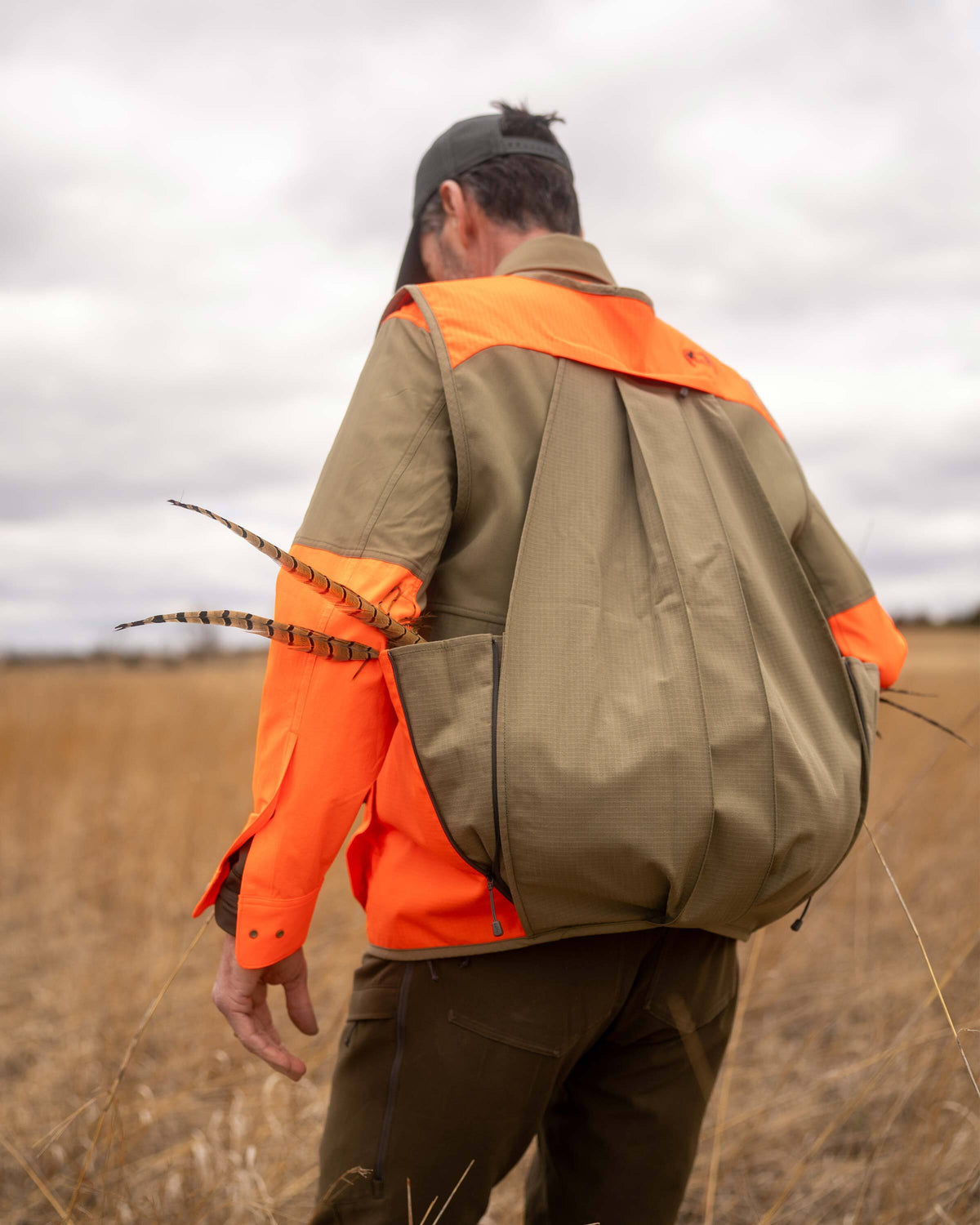 Back view of man wearing Core Upland Vest in Khaki/Blaze Orange, highlighting the wrap-around bird bag with a pheasant tail visibly sticking out.
