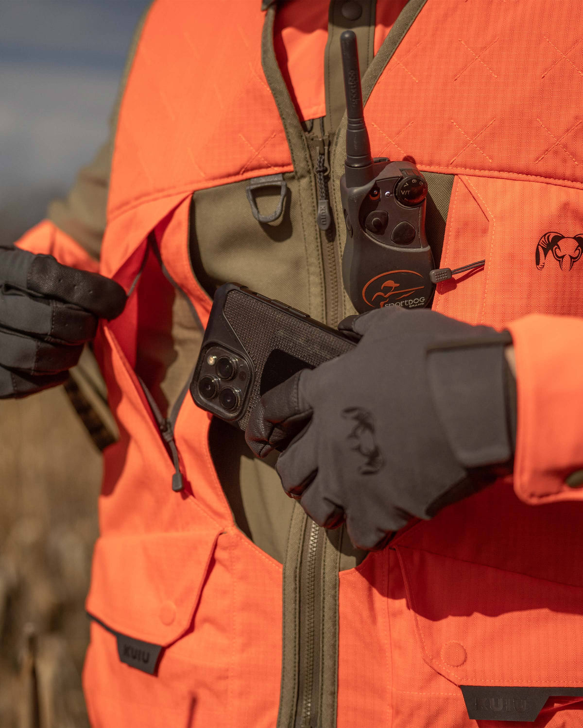 Man wearing Core Upland Vest in Khaki/Blaze Orange displaying a vertical zippered chest pocket on the right side with flap covers and storing his cell phone