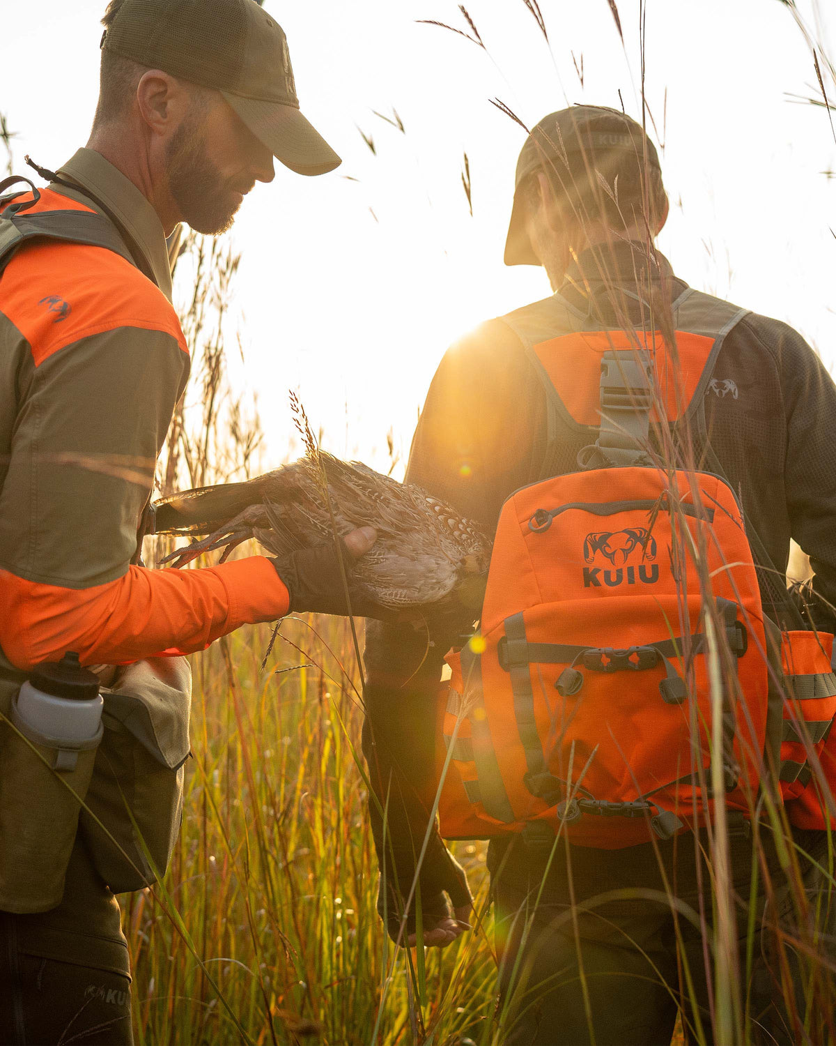Two hunters stand in a grassy field, one hunter holding a pheasant in his hands, preparing to place it into the pouch of the Upland Field Vest worn by the other hunt