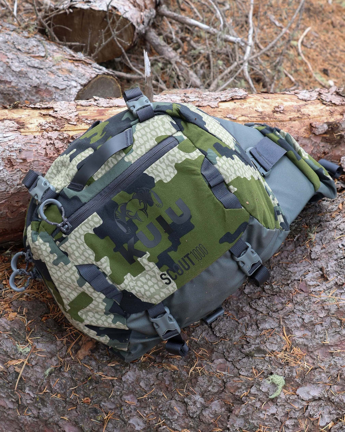 Front view of Scout 1000 in Verde Camouflage resting on the ground over a tree surface, surrounded by bark