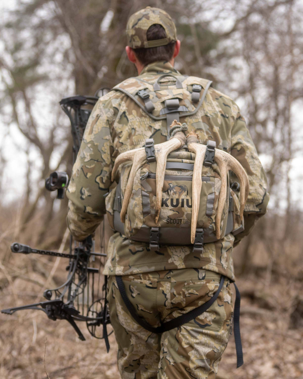 Whitetail hunter backpacking through a bare forested area, carrying a compound bow in the left hand and Scout 1000 in Valo Camouflage on his back with antlers attached