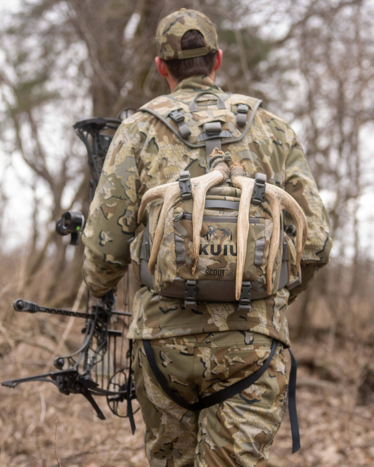Whitetail hunter backpacking through a bare forested area, carrying a compound bow in the left hand and Scout 1000 in Valo Camouflage on his back with antlers attached