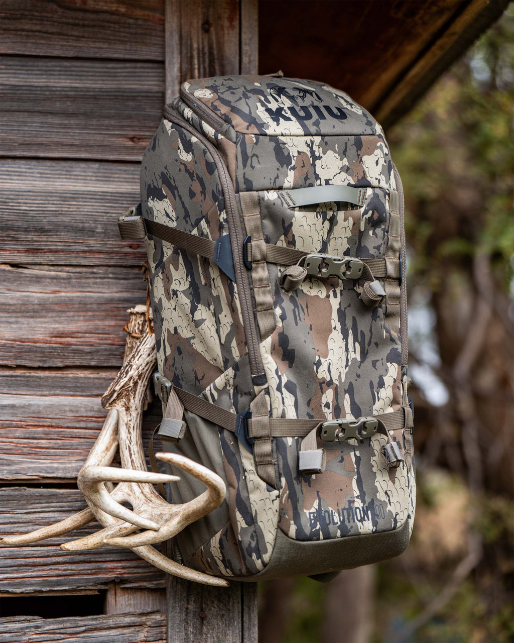 Evolution 1800 backpack in Veros Camouflage displayed hanging from a wooden post next to a set of deer antlers.