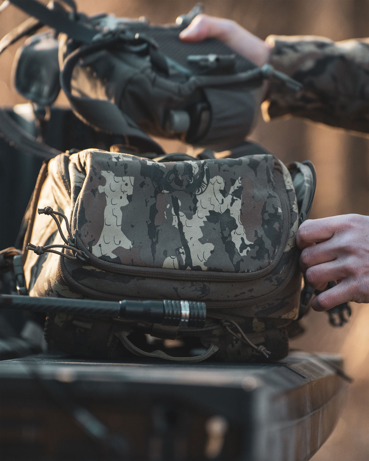 A close-up of Evolution 1800 backpack in Veros camouflage being handled by hands, with another bag in the background on a dark surface in an outdoor setting.
