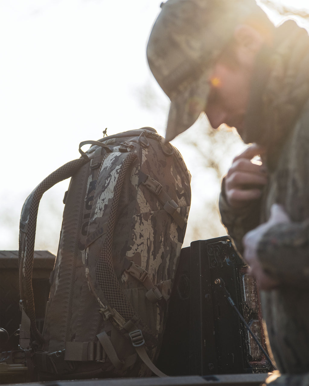 Evolution 1800 backpack in Veros Camoulfage sits in the bed of a truck next to a person wearing a matching Veros camouflage with bright backlighting.