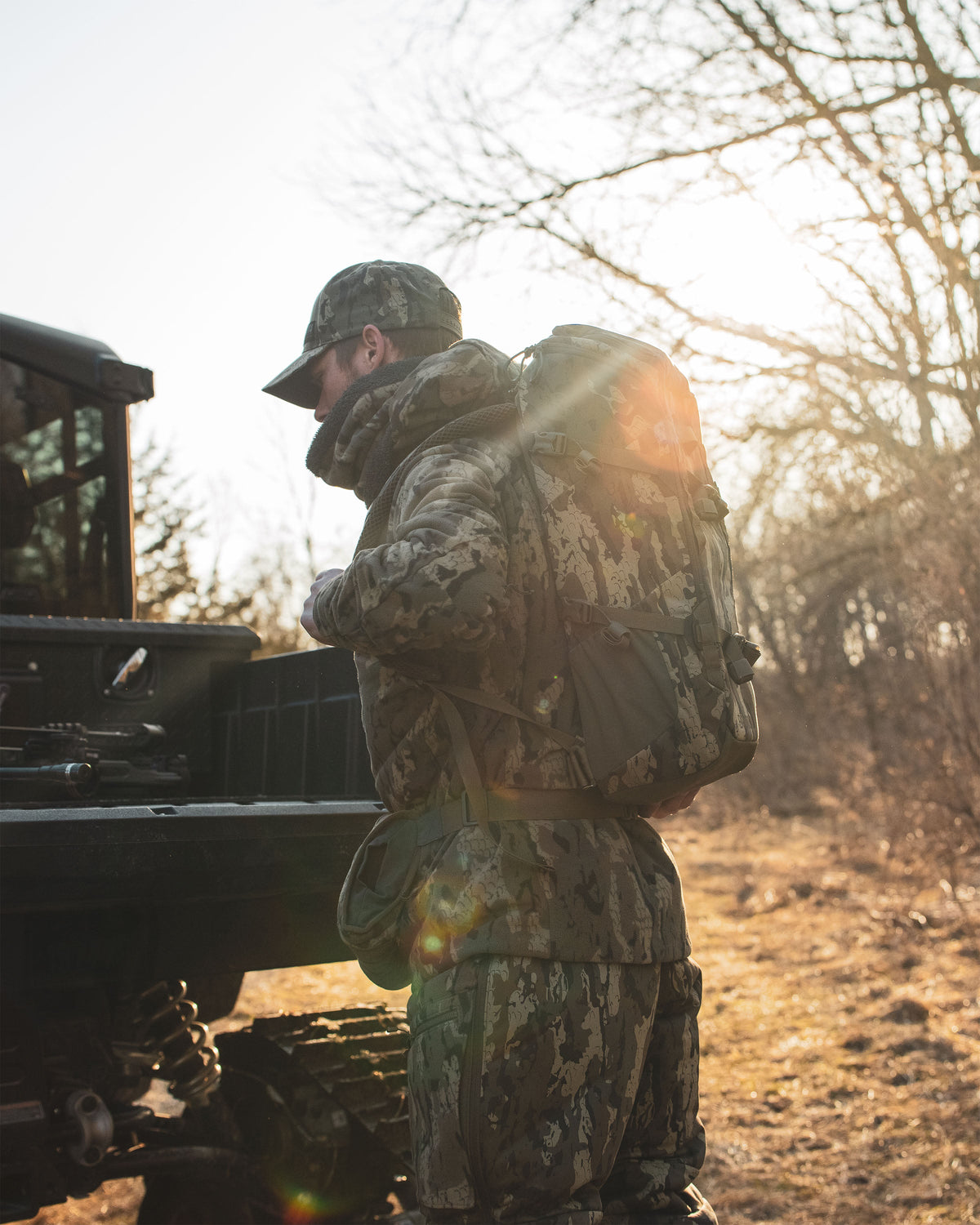 A person in full Veros camouflage outdoor gear, including a Evolution 1800 backpack in Veros Camouflage, stands beside a vehicle in a wooded, sunlit outdoor setting.