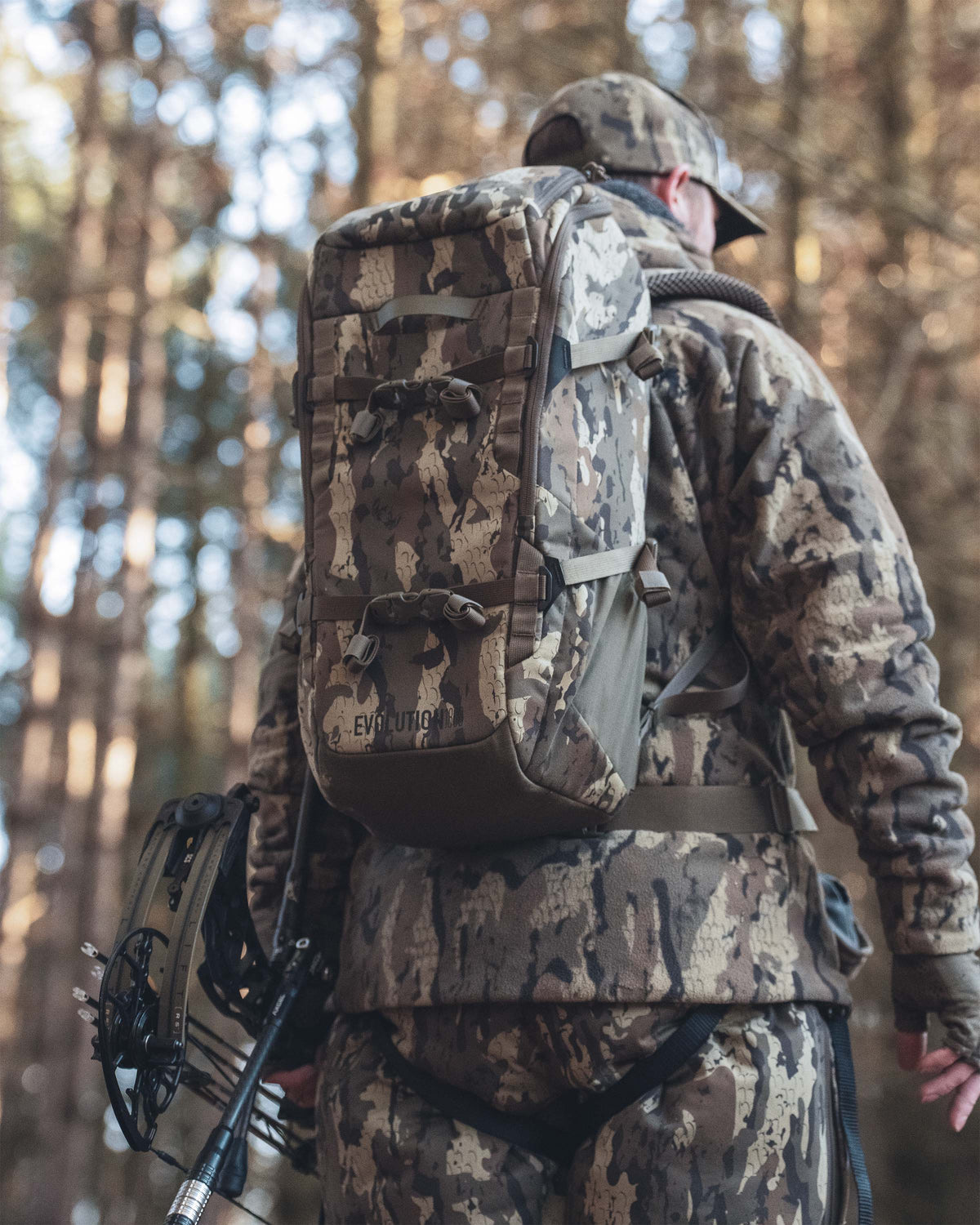 A person in full Veros camouflage hunting outfit seen from behind, wearing a Evolution 1800 Veros camouflage backpack outdoors in a forest.